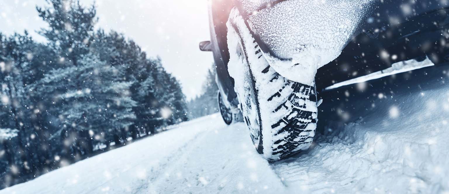 Car tire with snow tread driving on a snowy road with falling snow and pine trees in the background.