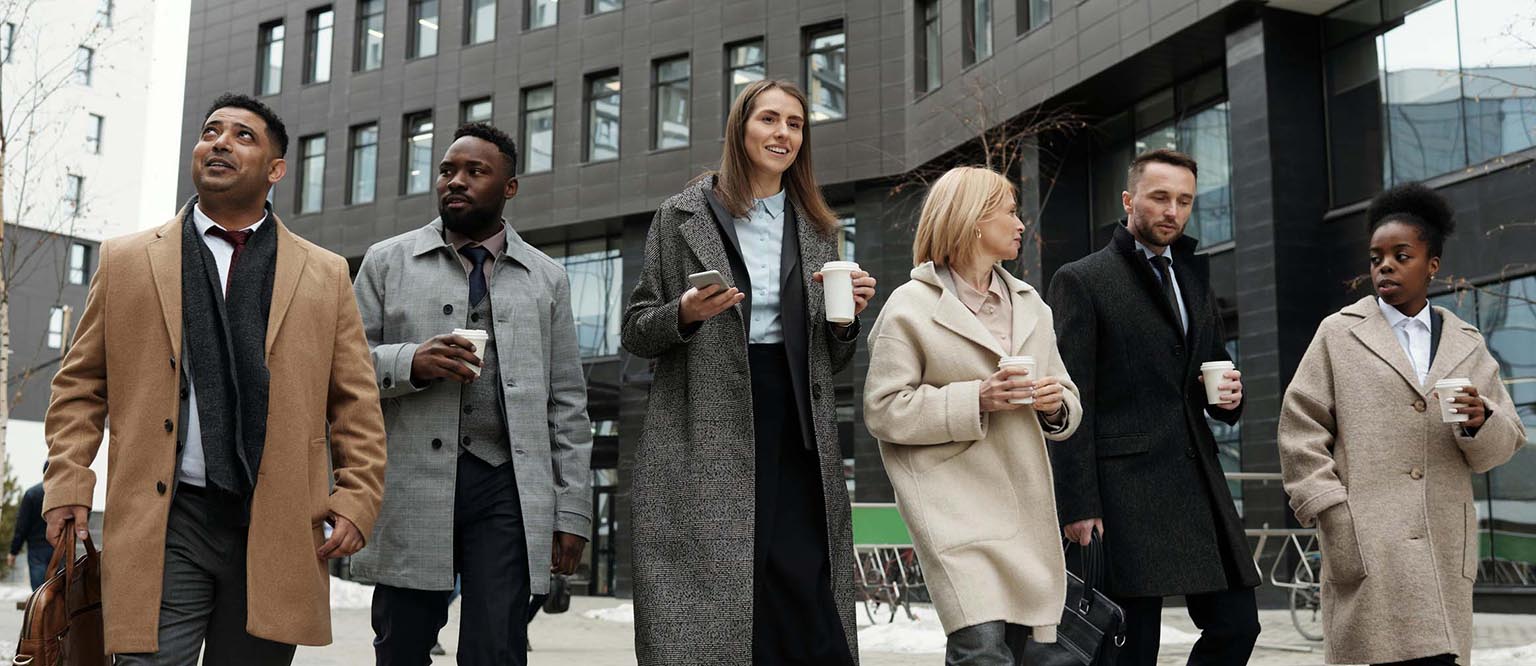 Group of six diverse business professionals walking outside an office building, dressed in winter coats and holding coffee cups, engaged in conversation.