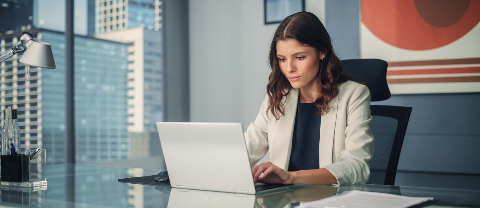 A woman in a white blazer works intently on a laptop at a sleek - https://ribeirodesousa.com/