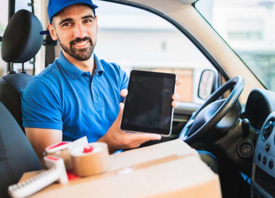 A smiling man inside a car, holding a tablet and looking at the camera.